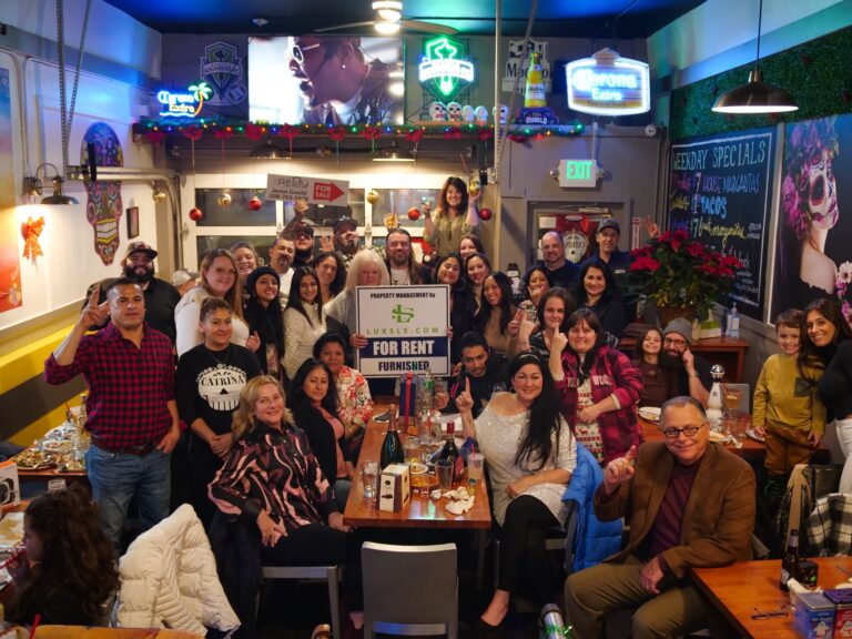 The Luxsle team at the 2022 holiday party, packed around the tables of a neighborhood restaurant with a Luxsle &lsquo;For Rent, Furnished&rsquo; sign in the middle