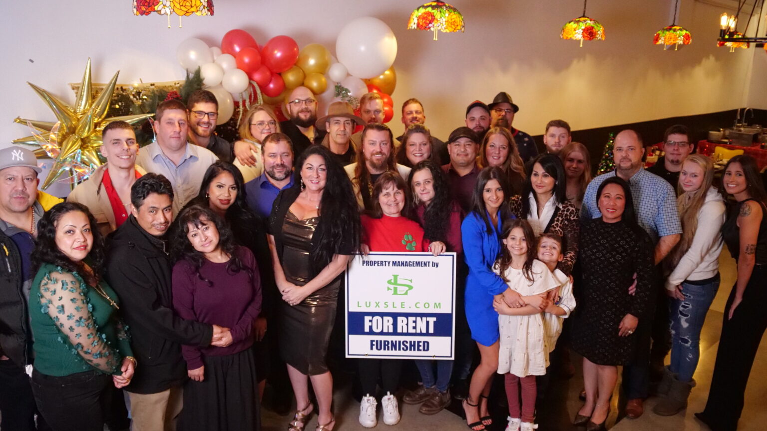 The Luxsle team at the 2023 holiday party, posed under a red and gold balloon arch around a Luxsle &lsquo;For Rent, Furnished&rsquo; sign