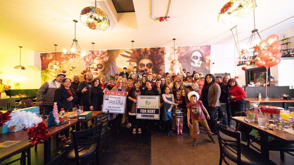 The Luxsle and Reluty team at the 2024 holiday party — around forty staff, partners and their children gathered in a restaurant, holding a Luxsle &lsquo;For Rent, Furnished&rsquo; sign and a Reluty &lsquo;For Sale&rsquo; sign