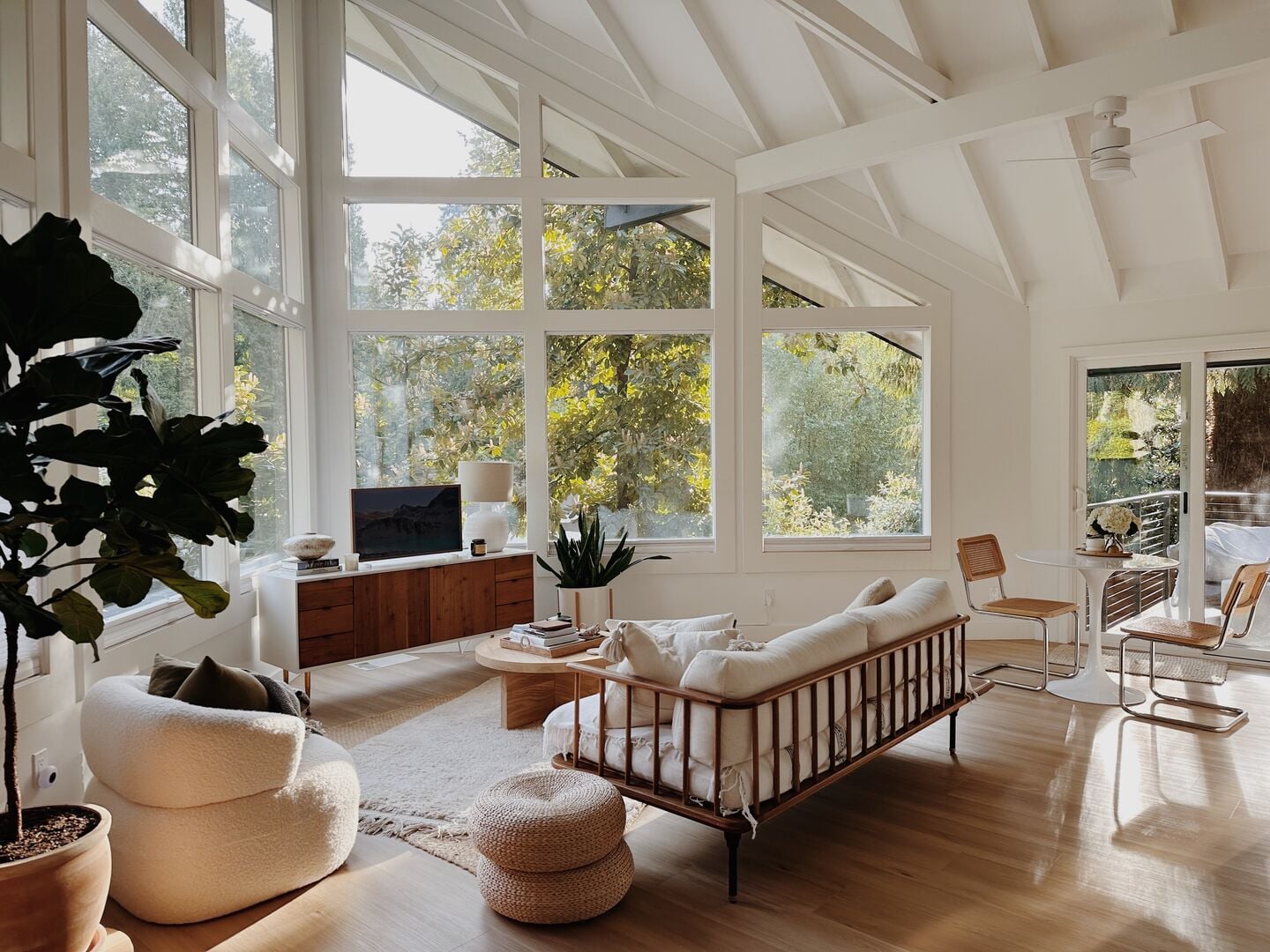 Sunlit living room under a vaulted white-beamed ceiling, with walls of tall windows onto trees, a cream sofa and a walnut sideboard