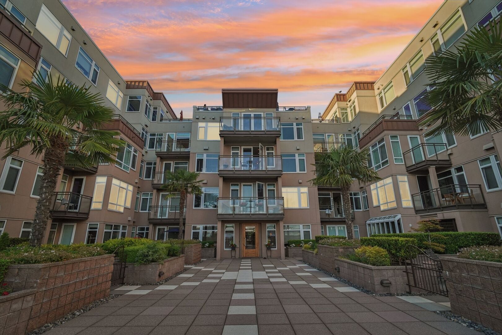 The courtyard of a four-storey condominium building at sunset, lined with palms and planters and lit from its windows