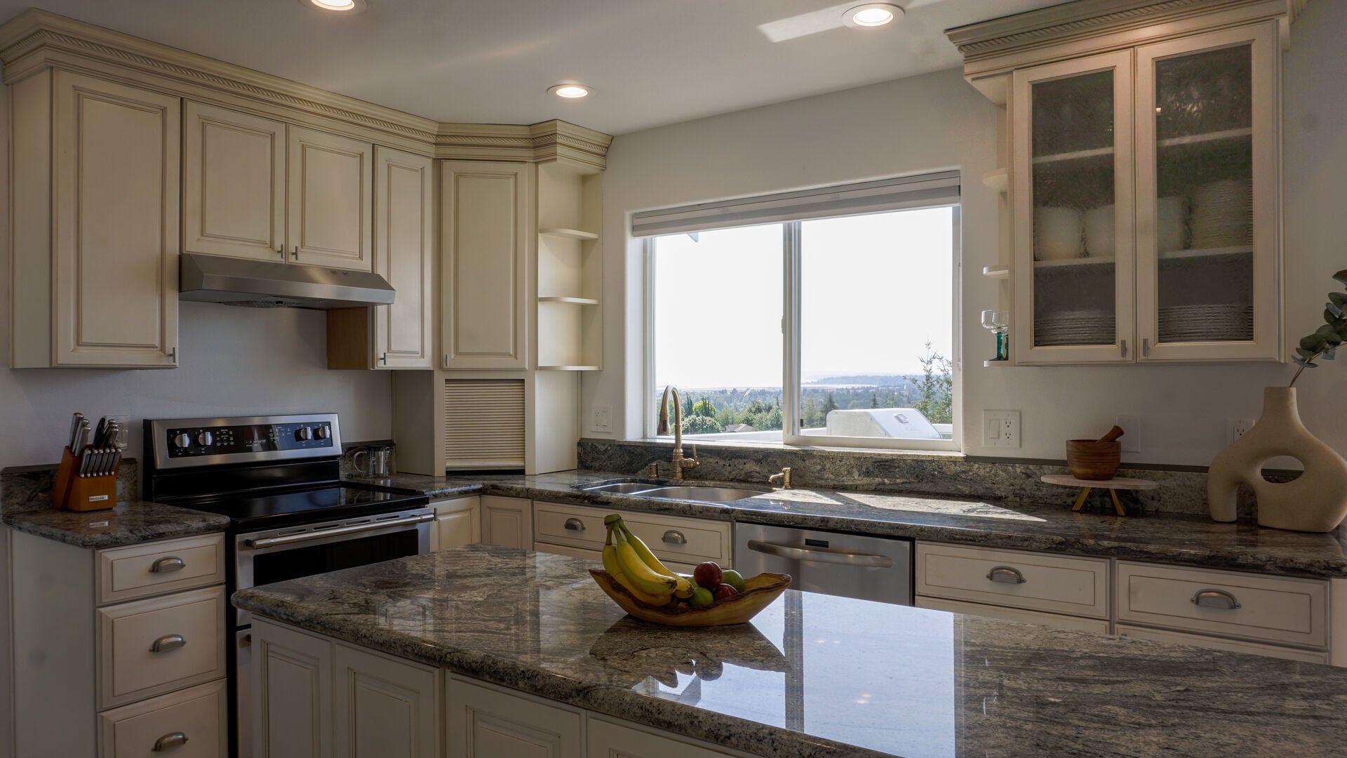Bright kitchen with cream cabinets, granite counters and an island, and a window looking out over trees toward the water